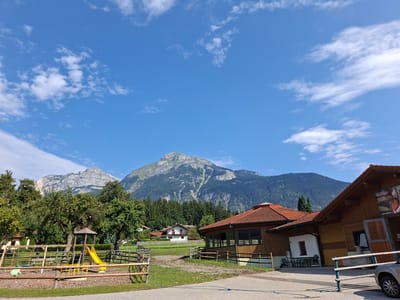 This is a horizontal, outdoor, medium-shot photograph taken on a sunny day in Münster, Austria. The scene depicts a rural setting with a playground and buildings against a backdrop of majestic mountains.
In the foreground, a wooden fence encloses a playground with a yellow slide, a wooden climbing structure, and a swing. To the right of the playground, a wooden building with a red-tiled roof and large windows is visible. A car is parked to the far right of the frame. Further back, behind the playground and the first building, are more wooden fences and green fields. Several trees, some laden with fruit, are scattered throughout the scene.
In the mid-ground, a cluster of houses with white and brown exteriors nestle amongst trees. Dominating the background is a vast, rugged mountain range. The lower slopes of the mountains are covered in dense, dark green conifer forests, while the upper reaches are characterized by rocky, grey cliffs and peaks. The sky above is a brilliant blue, sparsely dotted with white, fluffy clouds.
The overall impression is one of a peaceful, natural landscape complemented by recreational facilities and rustic architecture, characteristic of an Austrian rural resort or farm. The image captures a tranquil, idyllic moment in the Austrian countryside.