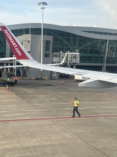An airport apron view shows a JetSMART plane wing with the company's website prominently displayed. A ground crew member, wearing a safety vest and holding a signal wand, stands beside the wing. The modern airport terminal, featuring a curved glass facade, is visible. The ground is marked with painted lines and tire tracks, illustrating an active airfield environment.