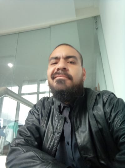 A medium, close-up shot of a man with brown skin, a full dark beard, and a partially shaved head. He is wearing a black leather jacket over a dark button-down shirt. The background features a wall of frosted glass panels and a white wall. The location appears to be indoors. The image does not provide sufficient information to confirm it was taken in Parroquia Guatire, Venezuela.