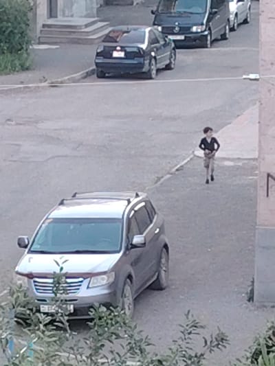 A young boy, wearing a dark long-sleeved shirt and light-colored shorts, walks across a paved area. He is in the mid-ground of the image, with several cars parked behind him and to his left. In the foreground, a silver SUV is parked, partially obscured by greenery. The scene appears to be an outdoor urban or semi-urban environment, possibly a street or parking lot, with buildings visible in the background. The lighting suggests it is daytime, with no strong shadows indicating a specific time of day. The weather appears to be overcast or neutral. The location is identified as the border of Gyumri-Akhuryan, Armenia. No visible text is discernable on the boy or the surrounding objects.