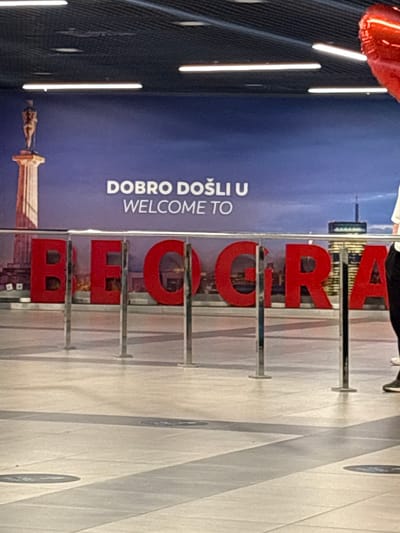 A red heart-shaped balloon is partially visible in the upper right corner of the image. In the background, a large blue mural displays the text "DOBRO DOŠLI U WELCOME TO" above a cityscape. Below the mural, large red letters spell out "BEOGRAD," with metal stanchions placed in front of them. The floor is tiled with a pattern of light and dark grey lines. A person wearing dark pants and a white shirt is visible standing to the right of the red letters, facing towards the left. The scene appears to be set in an indoor public space, possibly an airport or train station, in the city of Surčin, Serbia.