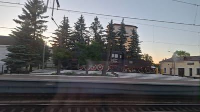 This image captures a scene at the Volkhov-Pribrezhnaya railway station in Volkhov, Russia. A dark, vintage steam locomotive, identified by the number "3'708-64" and Russian text, is on display on a raised platform. The locomotive is surrounded by several tall evergreen trees. Behind the locomotive, a multi-story building with a beige facade and a darker top section is visible. To the right of the locomotive, a small, yellow building with a red roof, possibly a station house or utility building, stands. The platform in the foreground is lined with white decorative elements. In the far background, the sky is clear with a pale gradient, suggesting either early morning or late evening. Railway tracks are visible in the foreground. No people are present in the image.