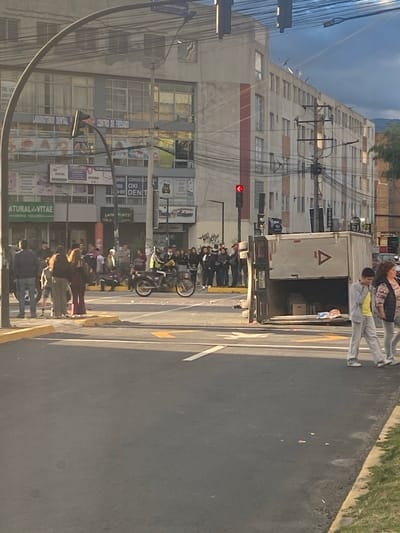 This image shows a street scene in Quito, Ecuador. A group of people are gathered on a sidewalk near a truck. The truck is white with a red triangle logo. A license plate on the truck reads "PBY-4309". Behind the people and truck is a tall, modern building with many windows. The building has signs on it, including "LABORATORIO DENTAL", "CENTRO DE FRESADO", "CONSULT", "MSD OXI DENT", and "ATURAL VITAE centro naturista". There are also traffic lights visible, with one showing red. The sky appears to be overcast. The time of day appears to be late afternoon or early evening, given the lighting. The overall scene is busy, with many people and vehicles present, suggesting an urban environment. The presence of medical and dental-related signage on the building indicates a commercial or medical district.