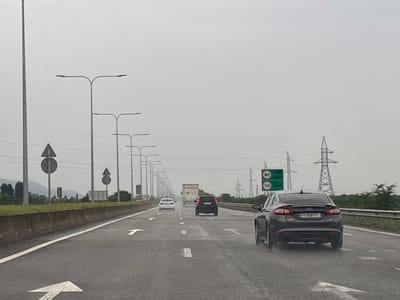 The image depicts a wet highway with multiple lanes of traffic under an overcast sky. In the foreground, a dark gray sedan is visible from the rear, driving on the right side of the road. The road surface is asphalt and appears damp, with lane markings including white arrows indicating direction. Several streetlights line the left side of the highway, and on the right side, there are power lines and signs. One sign shows speed limits of 80 and 110, with icons of cars. In the distance, a truck and other vehicles are visible, along with trees and hills on the horizon. The scene appears to be on a major road outside of urban development.