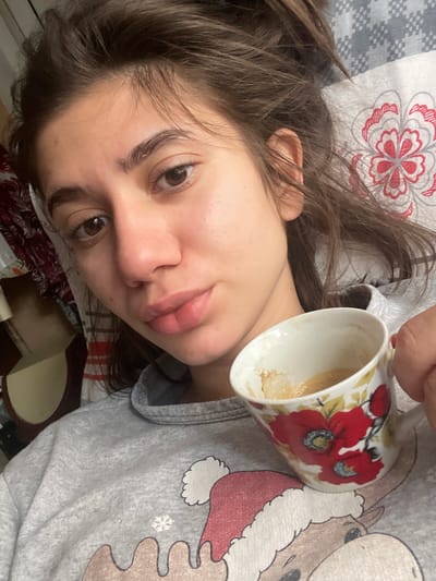 A woman is lying on a bed, holding a floral mug that appears to have coffee remnants inside. She is wearing a gray top with a cartoon design and is positioned at an angle. The bed has patterned linens. The location is Aprilovo, Bulgaria.
