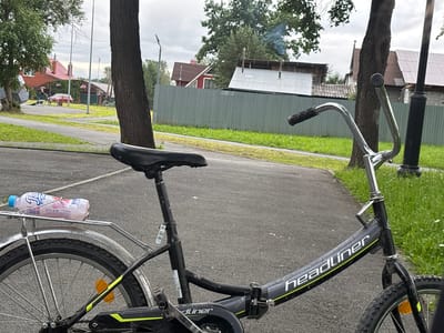 A medium shot captures a black bicycle parked on a paved path. The bicycle features a black seat, chrome handlebars, and a silver rack holding a partially filled pink plastic bottle. Visible text on the bicycle frame reads "headliner". In the background, a gray corrugated metal fence separates the path from a grassy area. Beyond the fence, several residential buildings with red and brown roofs are visible, along with trees. Smoke is seen rising from a chimney on one of the buildings, suggesting a cool or overcast day. The overall scene appears to be a quiet residential area in Nevyansk, Russia, likely in the late afternoon or early evening given the soft, diffused light. No people are actively engaged in activity, but the presence of parked bicycles and residential structures indicates this is a living space. The mood is peaceful and ordinary.