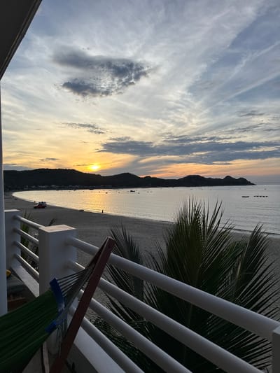 A wide-angle shot captures a tranquil sunset over a beach in Song Cau, Vietnam. The sky is a canvas of muted blues and grays, streaked with wispy white clouds, transitioning to soft oranges and yellows near the horizon where the sun is setting behind a dark, silhouetted mountain range. The calm sea reflects the sunset's glow, with small boats scattered on its surface. The sandy beach is mostly empty, with a few distant figures walking. In the foreground, a white railing frames the view. To the left, a green hammock hangs from a wooden frame, partially obscuring the railing and the beach. Palm fronds in the foreground add a tropical touch to the scene. A red vehicle with a white roof is parked on the sand near the water's edge.