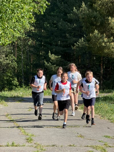 Five young people are standing in a line on a concrete path. They are all wearing white t-shirts with numbers on them, suggesting they are participating in a race or competition. The individual closest to the viewer has the number "4" on their shirt, and they are wearing grey shorts. The person behind them has the number "7" and is wearing black shorts with red accents on their t-shirt. The next person in line has the number "3" on their shirt and is also wearing black shorts. The person behind them has the number "9" on their shirt and is wearing black shorts. The last person in line has the number "6" on their shirt and is wearing black shorts. They all appear to be preparing to run or have just finished running, as they are standing close together in a line, possibly at a starting or finishing point. The concrete path is flanked by greenery, including trees and grass. The overall environment suggests a park or recreational area. The location seems to be a rural area within or near Oreshniki, Russia, as evidenced by the landscape and the style of infrastructure (path and buildings).