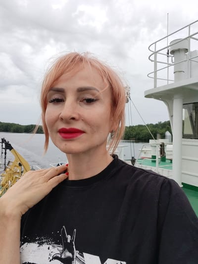 A selfie is taken by a woman on a boat, with her head and shoulders visible against a cloudy sky and a body of water. She has light pink, shoulder-length hair, pronounced eyebrows, and is wearing bright red lipstick. Her left hand is resting on her right shoulder, and her fingernails are painted red. She is wearing a black t-shirt with a graphic print, part of which is visible at the bottom of the frame.
In the background, a white structure of a boat or ship is visible, with railings and a white column. To the left, a yellow crane or piece of equipment is partially visible. Beyond that, trees and a body of water suggest a natural setting, likely on a river or lake, given the Novokemskii, Russia location context. The weather appears overcast, indicated by the cloudy sky. The woman's expression is neutral, and she is looking directly at the camera. No visible text is present.