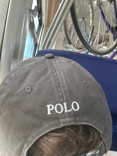The media file shows a close-up view of the back of a person's head wearing a dark grey baseball cap. The cap has white embroidered lettering that reads "POLO". The person's brown hair is visible peeking out from under the cap. In the background, out of focus, are parts of two bicycles, one with a purple frame and the other with a blue frame, and a portion of a blue padded object, possibly a seat or cushion. The setting appears to be indoors, with a metallic railing and a window or reflective surface in the lower portion of the image, suggesting a boat or a covered outdoor area. There are no explicit indicators of time of day or weather, but the lighting seems to be natural. The context places this scene in Salses-le-Château, France.