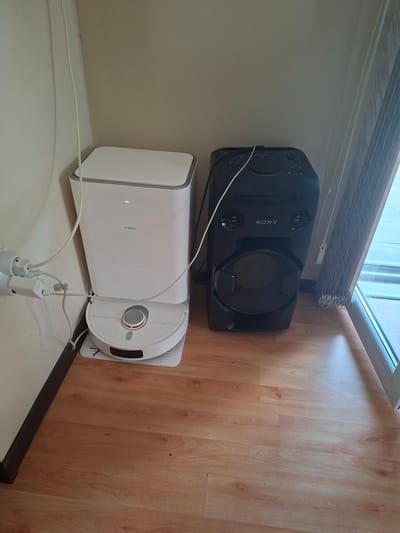 The image depicts a corner of a room with a light-colored wall and a wooden floor. On the floor, to the left, is a white Xiaomi brand robot vacuum cleaner docked on its charging base. The vacuum cleaner is white with a silver circular control panel on top. Adjacent to the robot vacuum, and to its right, is a tall black Sony brand stereo system. The stereo system has a large circular speaker at the bottom and two smaller speakers above, with various control buttons and a CD player slot on the front. Both the vacuum cleaner and the stereo system have white power cords connected. A white electrical adapter is plugged into the wall near the robot vacuum. To the right of the stereo system, a window is visible with vertical blinds partially drawn, revealing a bright outdoor scene. The overall scene is a tidy arrangement of electronic devices in a domestic setting, with no people present. The location is identified as Aksakovo, Bulgaria.