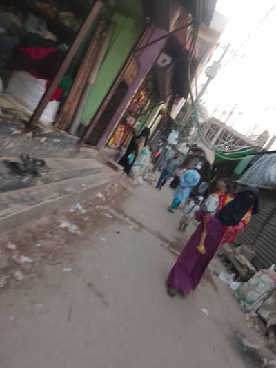 The image depicts a tilted, blurry view of a street scene in Tarabo, Bangladesh, during daylight. The street surface is light-colored and uneven, with scattered litter including plastic items and paper debris.
On the left side of the street, multi-level structures with distinct green and purple facades are visible. The ground level openings of these structures reveal bundled textiles and other merchandise, indicating commercial establishments. A set of concrete steps is also present on the lower left leading up from the street. Numerous overhead wires, forming a dense network, traverse the upper portion of the scene. On the right side, corrugated metal structures, consistent with market stalls or building exteriors, are partially visible.
In the foreground, a person wearing a long purple garment and a black head covering is seen carrying a small child on their left hip. The child is dressed in a light-colored top and yellow footwear. In the midground, several other individuals are present: one person is clad in a light blue long garment, another in a patterned light-colored garment, and a third person wears a darker top and blue trousers. A smaller child is also visible within this group. The scene portrays an active, populated urban environment.