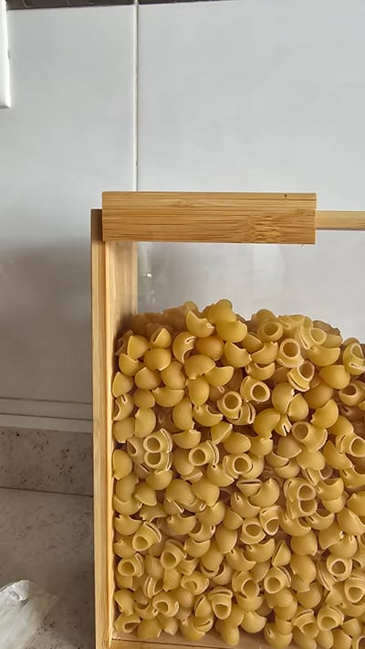 A close-up, eye-level shot shows a clear storage container filled with elbow macaroni. The container has a light brown wooden frame and a wooden lid. The background consists of a white tiled wall and a countertop. The image is focused on the pasta, with the wall and countertop slightly blurred. The location is not discernible from the image.