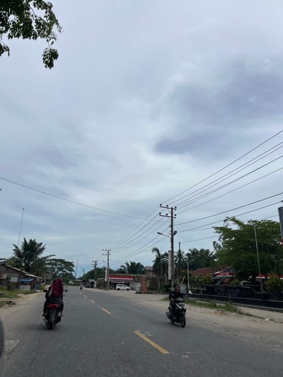 The image depicts a street scene in Seunebobaru, Indonesia, under an overcast sky. Two individuals are riding motorcycles in the same direction on a paved road with yellow lane markings. In the foreground, a person wearing a dark outfit and a maroon headscarf is riding a motorcycle. Further ahead, another person in a dark outfit is riding a black motorcycle. Along the sides of the road, there are various buildings, including small shops or houses with corrugated metal roofs, and some trees, including palm trees. Power lines are visible overhead, supported by utility poles. On the right side of the road, a building with signage that reads "SPBU 16.244.056" and "ACEH TARLING" is present. A white car is parked in front of one of the buildings. The overall environment suggests a rural or semi-urban setting.
