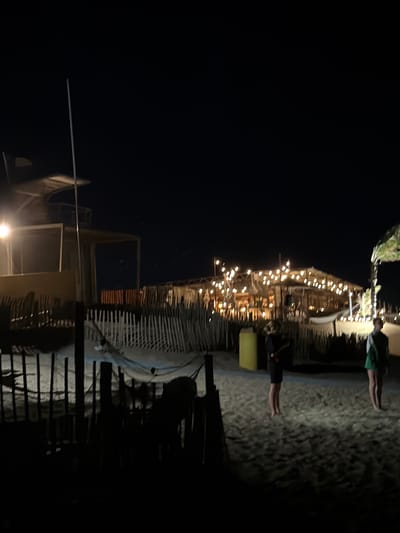 In Torreilles, France, on the beach at night, a group of people are gathered near a beach hut adorned with numerous lights. In the foreground, a white hammock is strung between two wooden posts, casting a faint shadow. Two individuals stand on the sand. One, a woman wearing a black dress, faces away from the camera. Another, a person wearing a green and white top and shorts, stands facing the camera. Behind them, a series of fences made of wooden planks extend across the sandy beach. A tall, thin white pole rises from the ground, extending out of the frame at the top. The scene is dimly lit, with the warm glow of the beach hut lights illuminating the immediate surroundings.
