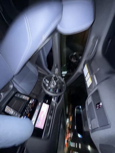 This media file presents a dynamic, motion-blurred view of the interior of a modern car, likely a Jaguar I-Pace, at night. The perspective is from the rear passenger side, looking towards the front of the vehicle.

The dominant subject in the foreground is the front passenger seat, upholstered in a dark, possibly blue-grey material with visible light-colored stitching, appearing blurred due to motion. Beyond it, the driver's seat, steering wheel, and dashboard are visible. The steering wheel, with its distinctive design and integrated controls, is motion-blurred but identifiable.

The center console features a contemporary design with two large digital screens. The upper screen displays what appears to be a navigation or infotainment interface with soft pink, purple, and blue hues. Below it, a second touchscreen controls climate and other vehicle functions, indicated by glowing orange and blue accents on buttons and dials.

Through the windshield and side windows, the external environment is a streaky blur of lights, signifying movement at night. Red tail lights, white headlights, and various streetlights or building illuminations create horizontal light trails against the dark background, suggesting the car is driving through an urban or suburban area such as Culver City, United States.

Overhead, the car's interior roof lining is visible, along with an overhead console containing interior lights and possibly controls for a sunroof. Sun visors are present, displaying unreadable warning labels. A rearview mirror is also visible, reflecting some of the exterior light trails. No people are distinctly visible in the frame, but the interior setup implies a driver and passenger.