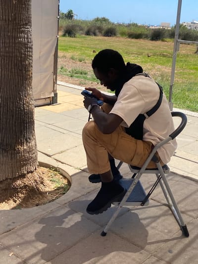 A dark-skinned man is seated on a folding metal chair, looking down at a black smartphone in his hands. He is wearing a light-colored t-shirt, tan pants, and black shoes. A dark bag strap is visible across his t-shirt. The man is sitting outdoors on a paved area with a textured brown tree trunk to his left. Beyond the paved area is a grassy field with some low bushes and trees in the background. The sky is clear and blue, suggesting a sunny day. The scene appears to be in a public outdoor space, possibly a park or recreational area, within the city of Conil De La Frontera, Spain.