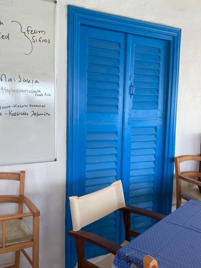 The image displays an interior view of what appears to be a dining establishment, characteristic of Greek architecture, located in Φάρος, Greece. A prominent, vibrant blue double door or window, featuring louvered panels, occupies the central and right portions of the frame, set into a white, textured wall. A dark handle is visible at the center seam of the blue doors. To the left, a white magnetic whiteboard is mounted on the wall, displaying handwritten text in Greek and English, indicative of a menu. Visible items include "from Sifnos", "Παϊδάκια" (Lamb chops), "Φαρόσουνα with fresh fish", "Κατσίκι κοκκινιστό" (Goat stewed in tomato sauce), and "Κατσίκι Λεμονάτο" (Lemon goat). In the immediate foreground, a wooden chair with a light cream-colored fabric backrest is positioned in front of the blue door. To the right of this chair, a table covered with a blue tablecloth patterned with white polka dots is partially visible. A second wooden chair with a light-colored seat is partially present in the bottom-left corner, and a third wooden chair is partially visible on the far right. The walls are uniformly white, providing a bright backdrop to the blue door and wooden furniture.