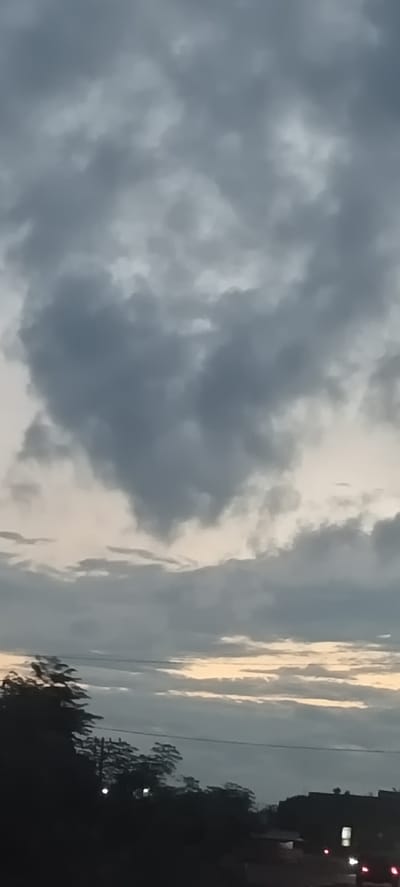 The image presents a twilight sky, dominated by dense, grey, and overcast clouds. A distinct heart-shaped cloud formation is clearly visible in the upper-middle portion of the frame. Below the heavy cloud cover, a strip of lighter sky peeks through near the horizon, exhibiting soft transitions from pale yellow to light orange, suggesting either sunrise or sunset.

The lower section of the image features a dark, silhouetted landscape. On the left, several trees with dense foliage are visible, while on the right, dark shapes that appear to be buildings or structures extend towards the horizon. A thin, horizontal line, likely a power line, cuts across the middle ground. Several small, bright points of light, consistent with artificial illumination from streetlights or vehicles, are discernible within the darkened foreground.

The scene conveys a calm and somewhat muted atmosphere, with the soft, diffused lighting characteristic of low-light conditions. The prominent heart-shaped cloud adds a unique and emotional detail to the cloudy sky. No human figures or discernible text are present. The setting, located in Meunasahmee, Indonesia, appears to be an outdoor scene looking towards a distant settlement or road during the transitional period of day and night.