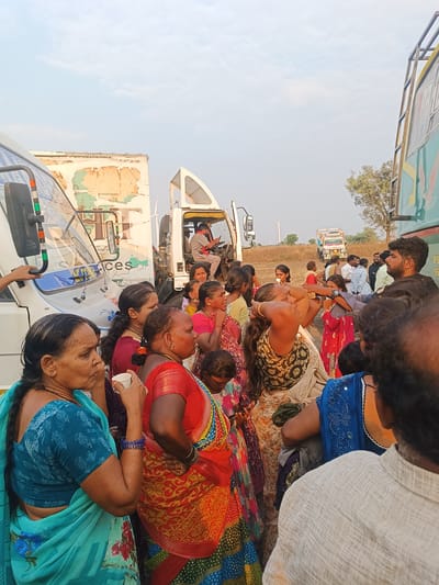 This image depicts a daytime gathering of approximately 20-30 individuals, predominantly adult women dressed in traditional Indian attire, in a dry, open, rural environment in Kottalapalli, India. The scene shows multiple large vehicles stopped on what appears to be a dirt path or roadside.
On the far left, the front section of a white bus or large van is visible. Behind it, a larger white truck displays weathered signage with partially visible text including "HYDERABAD", "CHENNAI", and "Services". To the right of this, the cab of another white truck is seen, with a man inside operating a mobile phone. On the far right, the side of a green and yellow bus is partially visible. In the distant background, another colorful truck is stopped on a dirt track, and a wind turbine is visible against the clear sky.
People are clustered between and around these vehicles. In the left foreground, a woman in a turquoise saree holds a small white cup to her mouth. Another woman in a red and gold saree stands beside her. A bearded man with dark hair is visible on the far right, facing towards the left side of the frame. The individuals appear to be standing, waiting, or conversing in small groups. The overall impression is that of a roadside stop or a temporary halt during travel.