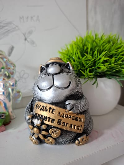 A close-up, eye-level shot focuses on a figurine of a grey cat wearing a golden hat, holding a golden sign with Russian text. The cat is depicted with a smiling expression, its paws resting on the sign. Scattered around the cat's paws are golden objects resembling coffee beans or small stones. To the left of the cat, partially visible, is another figurine, possibly a person, with a shiny, iridescent finish. In the background, a white pot holds a vibrant green artificial plant. The surface the figurines are placed on is white and appears to be a shelf. Faintly visible on the wall behind the figurines is a sketch that includes the Russian word "МУКА" (flour) and a drawing of a butterfly. The overall lighting is soft, with the focus clearly on the cat figurine. The scene does not contain any specific information to indicate its location in Korenovsk, Russia.