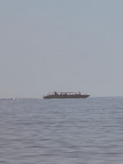 A long, narrow passenger boat with a dark hull and a light-colored superstructure is navigating on a calm, gray body of water. The boat appears to be carrying passengers. The sky is a uniform, pale gray, suggesting overcast conditions or a hazy atmosphere. The scene is likely a coastal or large lake environment. The image has a slight grainy texture, indicative of a photograph taken at a distance or with limited light. The boat is positioned roughly in the center of the frame, with the water occupying the lower half and the sky the upper half. There is a small, bright white object visible on the water to the left of the boat.