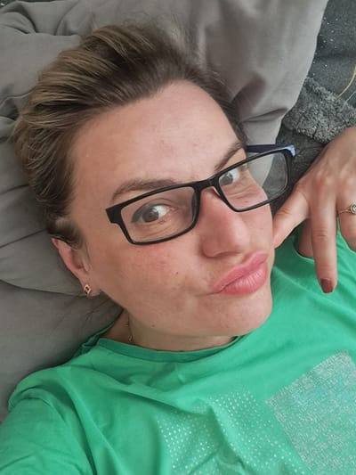 A middle-aged woman lies on a gray surface, possibly a bed or sofa, wearing a green shirt with a faded white pattern. She is positioned with her head facing the viewer and her body angled to the right. The woman is wearing black-framed eyeglasses, and her left hand is raised near her forehead, displaying a ring on her finger. Her hair is blonde with darker roots. She is wearing lipstick. A gold chain is visible around her neck. The photo is taken from a slightly overhead perspective. The location is not discernible from the image.