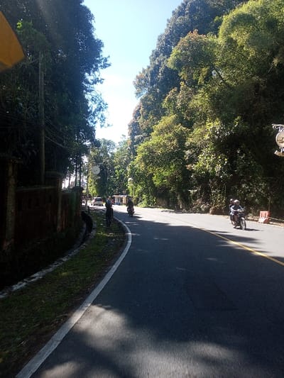The image depicts a paved road in Ka Thaung (Upper), Myanmar, curving gently to the right under bright sunlight and a clear blue sky. Dense, green tropical foliage lines both sides of the road, with trees forming a canopy overhead. The asphalt road features a solid white line marking its left edge and a double yellow line visible in the distance, indicating traffic flow in both directions.
On the left side of the road, a low, dark-colored wall, possibly brick or concrete, runs parallel to the asphalt, separated by a narrow strip of grass and a drainage ditch. A utility pole with multiple overhead wires is positioned behind this wall. Further along the left side, a white van or SUV is stopped or parked on the shoulder, with two individuals standing nearby on the roadside. Beyond this, two small structures with blue roofs are visible. A street light pole equipped with multiple lamp fixtures and a circular white dish-like object is also present on the left.
Three motorcycles are visible on the road. One motorcyclist, wearing a light-colored helmet and dark clothing, travels away from the viewer in the left lane. A second helmeted motorcyclist follows further ahead in the same direction. A third motorcyclist, also wearing a light-colored helmet and dark clothing, is approaching from the distance on the right side of the road. Distinct shadows from the overhead trees are cast across the road surface. The right side of the road is bordered by a steep, densely forested hillside.
