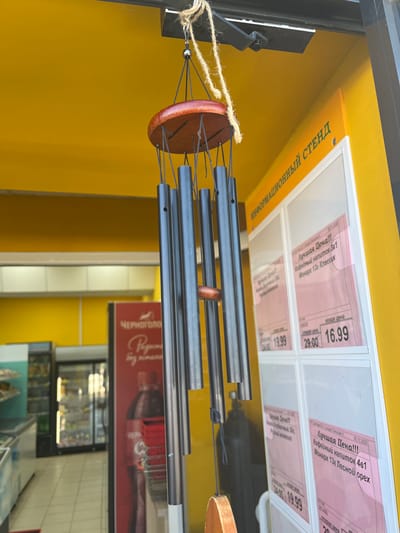 The scene depicts the entrance area of a retail establishment in the city of Сундур, Russia. A wind chime, featuring a round reddish-brown wooden top, eight dark metallic tubes of varying lengths, and a small central wooden clapper, hangs suspended by twine and a metal hook from a black metal bracket attached to a yellow ceiling or awning structure.
To the right of the wind chime, an "ИНФОРМАЦИОННЫЙ СТЕНД" (Information Stand) is affixed to a bright yellow wall. This stand contains multiple clear plastic pockets displaying pink price labels with Russian text. One label prominently reads "Лучшая Цена!!!" (Best Price!!!) for "Кофейный напиток 3в1 Монарх 13г Классик" (Coffee drink 3-in-1 Monarch 13g Classic), showing a crossed-out old price of 29.00 and a new price of 16.99. Another visible label pertains to "Кофейный напиток 4в1 Монарх 13г Лесной орех" (Coffee drink 4-in-1 Monarch 13g Forest Nut) with a new price of 19.99.
Through an open doorway or archway to the left, the interior of the store is visible, indicating a retail environment. This interior features multiple commercial refrigerators or freezers with glass doors, displaying products. A large red vertical sign or banner for the "ЧЕРНОГОЛОВКА" (Chernogolovka) brand, featuring a dark beverage bottle, is visible within this interior. The floor inside the establishment appears to be tiled.