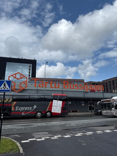 A vertically oriented image shows the Tartu Bussijaam (Tartu Bus Station) under a partly cloudy sky. Below the station's sign, a black and red "Express" bus is parked. A pedestrian crossing sign is visible in the lower left corner. The bus has a paper attached displaying a list of destinations, including "FRANCE", "BELGIUM", and "MOSCOW". A second bus, heavily covered in stickers, is parked further down the street.
