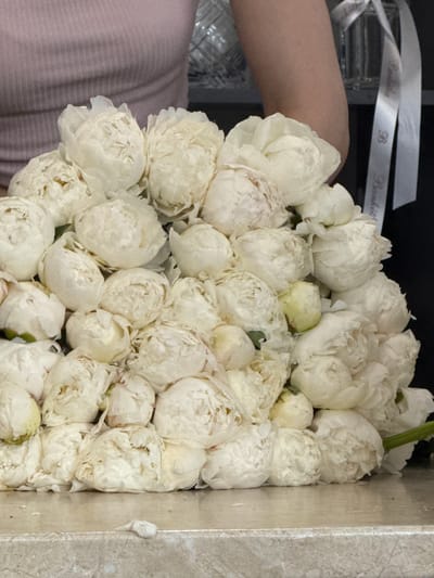 A close-up shot shows a large bouquet of white peonies, with some buds and fully bloomed flowers. The peonies are densely packed and arranged on a light-colored, textured surface that appears to be a marble countertop. In the background, a person is partially visible wearing a light pink, ribbed top. To the right of the bouquet, a white ribbon with dark text and a "B" logo is draped over a dark surface, suggesting a gift or floral arrangement service. The overall scene is bright and the focus is on the texture and volume of the flowers. The specific location in Assinovskaya, Russia is not discernible from the image.