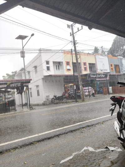 The image depicts a street scene in Paloh Teungoh, Indonesia, during a period of heavy rainfall, evidenced by visible rain streaks and saturated asphalt pavement. In the immediate foreground on the right, the rear portion of a dark-colored motorcycle with a chrome exhaust is partially visible, parked on wet ground containing white debris. The vantage point is from under a dark, overhanging structure.
Across the street, a row of two-story shophouses is visible, featuring various facade colors including white, yellow, and grey, each equipped with red-colored awnings over their ground-floor entrances. A prominent, heavily wired utility pole stands centrally in front of these buildings, with a solar-powered streetlight positioned to its left.
Under the awning of a shop identified as "Vanya Emil Shop," two individuals are observed: one person wearing a pink headscarf is seated on a motorcycle equipped with a sidecar, and another individual stands nearby. To the left, two additional motorcycles, one also featuring a sidecar, are parked on the sidewalk in front of a white building that displays a partially legible "NATION HOLD" sign under its awning.