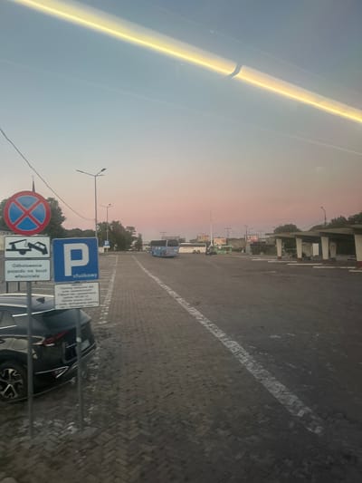 The image depicts a parking lot at the Lublin Główny bus station in Lublin, Poland. The sky displays a gradient of pale blue to soft pink, indicative of either dawn or dusk. In the foreground, to the left, a black SUV is parked. Next to it are several Polish traffic signs. One circular sign with a red border and a blue background with a red cross signifies "No Stopping." Below this is a rectangular sign with an image of a tow truck and text in Polish, indicating "Vehicle towed at owner's expense." A large blue square sign with a white "P" on it, also in Polish, indicates "Parking, service." Another sign below it provides parking regulations in Polish. The parking lot surface is paved with cobblestones. In the background, several buses are visible, including a blue intercity bus. A bus shelter with white pillars and a dark roof is present on the right side of the frame, with some people seated on benches beneath it. Streetlights are positioned throughout the parking area. The overall scene suggests a functional transportation hub during twilight hours.