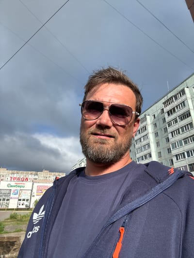 This is a selfie taken by a middle-aged man with short, tousled brown hair and a stubble beard. He is wearing sunglasses with reflective lenses and a dark blue hooded jacket with an orange zipper pull. The jacket appears to be from the brand Adidas, as indicated by the logo visible on the sleeve.

The man is outdoors, and the background suggests an urban setting in Usinsk, Russia. To his left, a tall, somewhat weathered apartment building with numerous windows is visible, extending upwards and out of frame. To his right, another building with signage is partially visible. One sign clearly reads "ОТРАДА" (Otrada) in large red letters, and below it, "Самобери" (Samobere). Other signs are also present, one with "10%".

The sky is cloudy and overcast, suggesting either early morning or late afternoon. The overall lighting is soft, with some areas illuminated by sunlight. The man's expression is neutral, and he seems to be looking directly at the camera. The angle of the shot suggests he is looking up, perhaps from a balcony or a lower vantage point.