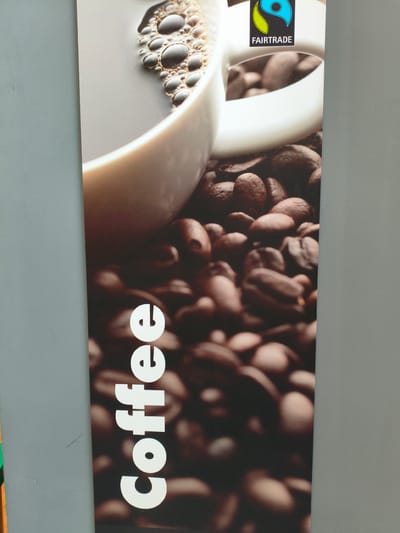 This media file is a vertical, close-up photograph of a poster or advertisement. The poster features a large, out-of-focus image of coffee beans, with a white coffee cup and some coffee with foam visible at the top. A Fairtrade logo is also present on the cup. The word "Coffee" is printed vertically in large, white, sans-serif font along the left side of the poster. The background of the image is a plain, textured gray wall, suggesting the poster is displayed indoors, possibly in a shop or cafe. The lighting appears to be even, indicating artificial light. There is no indication of time of day, weather, or specific emotions. The only text visible is the word "Coffee" and the "FAIRTRADE" logo.