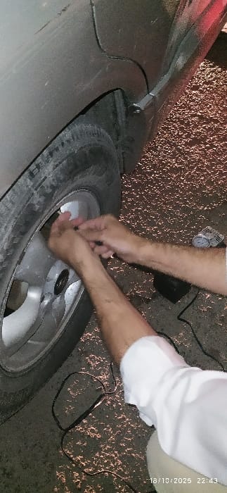 The image, captured at 22:43 on October 18, 2025, shows a close-up, low-angle perspective of an individual interacting with a vehicle's tire in Al-Kiswa, Syria, under artificial nighttime illumination.

The upper left portion of the frame displays a section of a gray or light brown sedan's rear quarter panel, exhibiting minor surface scratches. Below this, a dirty black tire is mounted on a silver-colored five-spoke alloy wheel.

The central and lower sections prominently feature the forearms and hands of an individual, dressed in a white long-sleeved shirt and light-colored trousers. The person is stooping or kneeling, with their hands positioned at the tire's valve stem, actively engaging in the process of connecting or disconnecting an air hose or adapter.

On the dark, textured ground, which appears to be asphalt with reflective speckles, a compact black portable electric tire inflator is visible, accompanied by a pressure gauge and a black electrical cable extending from it. No other individuals or significant environmental features are discernible within the cropped view.