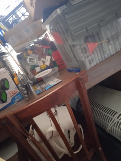 A wooden chair is in the foreground, with its backrest and seat visible. Behind the chair, a brown wooden table is laden with various objects. To the left, a toy race car in a blue and white box with green wheels is prominent. Next to it, a silver toy gun with a yellow accent is positioned. A clear plastic bin filled with colorful building blocks, including white window pieces and red bricks, sits on the table. A small white plastic container with a lid is also visible on top of the building blocks. Further to the right, a large clear plastic storage bin contains more items and two small red flags on sticks. A blue marker lies on the table near the bin. In the background, a white electric fan with a gridded cover is partially visible, and further back, a cardboard box with "R R" branding is stacked on another box. The scene appears to be indoors, possibly in a residential setting. There are no people visible in the image. The lighting suggests it is daytime, with light entering from the left.
