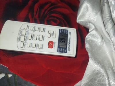 A remote control for an air conditioner is positioned on a red textured surface. The remote is white and features numerous buttons with labels such as "SET TEMPERATURE", "MODE", "SLEEP", "SWING", and "ECO". A digital display on the remote shows "86" with other smaller indicators. To the right of the remote, a white fabric with a patterned texture is draped. The red surface appears to be fabric as well, possibly a tablecloth or bedspread, with a pattern of dark red roses. The lighting is somewhat dim, casting subtle shadows. There is no indication of people or any activity taking place. The scene appears to be indoors, likely in a residential setting. No specific location in Tursunzoda, Tajikistan can be determined from the image.