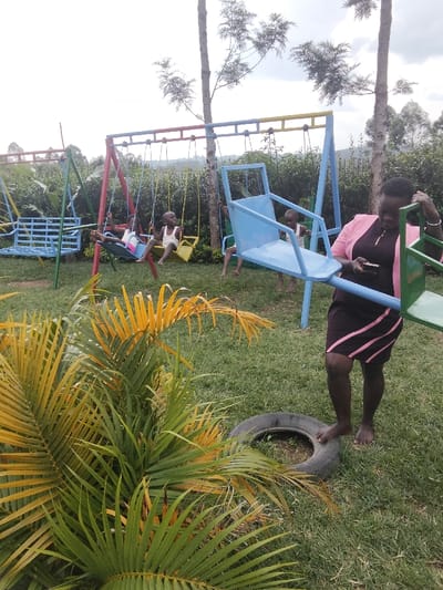 The image depicts an outdoor playground in Malaha Isongo Makunga ward, Kenya. The scene features a grassy ground cover with several pieces of metal playground equipment and natural foliage under an overcast sky.
In the foreground, a large plant with green and yellow palm-like fronds is positioned on the left. An adult female stands barefoot on the right side of the frame, wearing a dark dress with pink horizontal stripes on the lower half and a light pink long-sleeved jacket. She holds a light-colored mobile phone in her left hand, looking downwards, with her right hand near the frame of a blue metal seesaw. An old, black vehicle tire lies on the grass near her right foot.
In the background, multiple children are present on various playground structures. A light blue metal swing set with a bench-style seat is visible on the far left. Centrally, a multi-colored swing set (red and blue frame with yellow seats) supports three children: one on a blue swing, and two on yellow swings. An additional child is partially visible standing near the blue seesaw. The background consists of dense green foliage and several tall, slender trees.