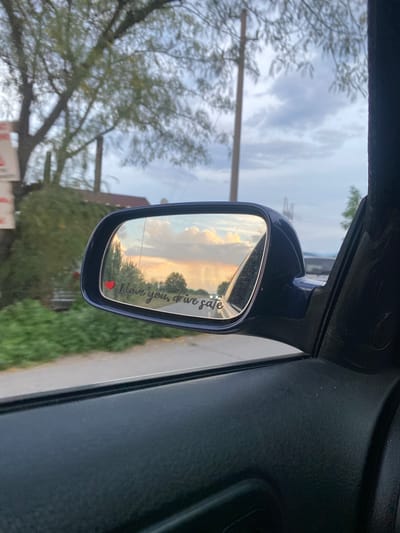 The image depicts a close-up view of a car's side mirror, reflecting a dusky sky with soft, warm clouds. A small red heart icon is visible on the mirror's surface, to the left of the text "I love you, drive safe" written in a cursive font. The reflection in the mirror shows a road with a few distant lights, trees, and power lines. The surrounding environment visible outside the car window includes lush green foliage and trees against a backdrop of a cloudy sky. The car's interior, specifically the dark-colored door panel, is also partially visible in the foreground. The scene is likely captured from inside a vehicle.