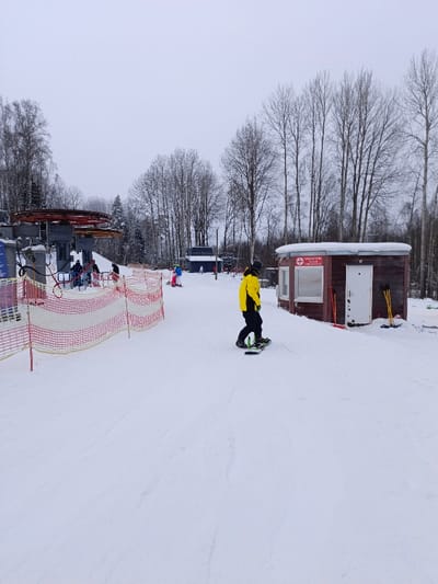 The image captures a snowy winter scene at a ski resort under an overcast sky. The ground is covered in white snow, showing various tracks and patterns from skiing and snowboarding activities.
In the immediate foreground and midground, a person wearing a bright yellow jacket and black pants is standing on a snowboard, facing slightly to the right and away from the camera. The snowboard is green and black. To their left, a red and white safety net or fence stretches across the snow, acting as a boundary.
To the right of the snowboarder, a small, rustic wooden building with a rounded roof is visible. The building is brown with white trim around its windows and a white door. A red sign with a white cross, the universal symbol for first aid, is prominently displayed above one of the windows. Below this symbol, Cyrillic text is visible, which reads "СПАСАТЕЛИ" (Rescuers) and "ПУНКТ ПОМОЩИ" (Aid Post), along with a website address "WWW.PRUDKI.RU". Leaning against the side of this building are several red shovels and yellow poles or brooms.
Further into the scene, to the left, a large ski lift mechanism with grey support structures and red operational components is visible. Several individuals are gathered near the base of this lift, some appearing to be waiting in line or preparing to use it. Another person, dressed in a blue jacket and yellow pants, is seen on skis further back in the midground, near a smaller, darker building which is partially obscured by trees.
The background is dominated by a dense forest of tall, bare deciduous trees, characteristic of a winter landscape. A few evergreen trees can also be seen interspersed among them. The sky is a uniform light grey, suggesting an overcast day. The overall impression is that of a functional, active ski area during winter.