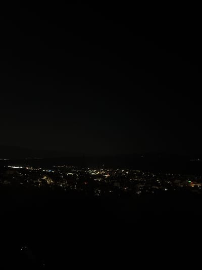 A night aerial view captures the city of Polienitsa, Bulgaria, illuminated by countless lights. The urban sprawl stretches across the landscape, a dense tapestry of glowing dots against the dark canvas of the night. Distant lights suggest a body of water or a vast expanse beyond the city limits. The foreground is dominated by darkness, with only faint, scattered lights hinting at the terrain or structures below. The scene is devoid of any human figures or discernible objects beyond the urban infrastructure.