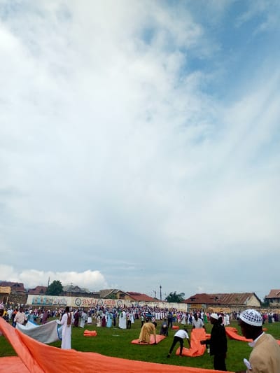 The image shows a large gathering of people in a grassy field in Beni, Democratic Republic of Congo, located at coordinates (latitude: 0.489, longitude: 29.463). The sky is bright blue with white puffy clouds. People are dressed in various clothing, including some wearing white robes and hats. Several people are bending over, folding a large orange tarp. In the background, there are a number of buildings with roofs, some with a red tile pattern, and a sign with the letters "BNT. ASBL" visible.  A large crowd of people are gathered in the distance.  The image was likely taken during an outdoor event.  The individuals are all adults, with some carrying items, including a few holding bags. There are no children visible in the photo.  There are no other objects of note besides the large orange tarp.  It appears to be a warm, sunny day.  The image was likely taken during the day as shadows are visible.  The grass in the foreground is green.