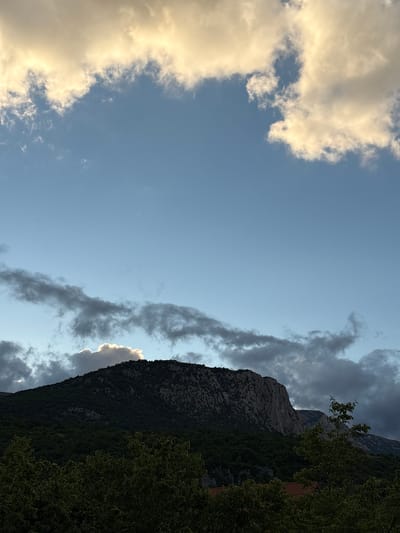 The image shows a mountainous landscape under a partly cloudy sky. The sky is blue, with large, fluffy clouds, some of which are tinged with yellow and white from the sunlight. Below the sky, a dark, forested mountain range dominates the scene. The peak of one mountain is illuminated by sunlight breaking through a cloud. In the foreground, the tops of dark green trees are visible. The location is identified as Malyi Mayak, Ukraine. There are no people or animals present in the image. The scene appears to be a natural landscape, possibly captured during a sunset or sunrise due to the lighting on the clouds.