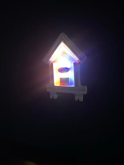 A translucent, miniature, house-shaped object, approximately 15-20 cm in height, is depicted against a uniformly dark, black background. The object is illuminated from within, emitting a multi-colored spectrum of light including red, orange, yellow, green, blue, and violet, which glows brightly and diffuses slightly into the surrounding darkness. Structurally, it features a clear, pointed triangular roof and a rectangular body resting on a base with four small, splayed legs. Inside the rectangular section, a standard Type G electrical socket is visible, positioned below an oval-shaped feature. No individuals, specific actions, or other discernible objects are present in the frame. The scene is observed within the city of Igbessa, Nigeria.