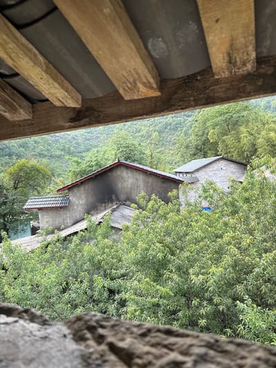 View from an elevated position, possibly a building, looking down upon a rural landscape in Sà Phìn Commune, Vietnam. The image contains:
1. **Built Environment:** Two visible concrete buildings with sloping roofs. One has a red trim along the roof edge. One has multiple windows visible.
2. **Natural Environment:** Lush green foliage partially obscures the buildings. A hillside or valley with dense vegetation is visible.
3. **Foreground Obstructions:** Elements from the viewing location are present: Wooden slats and curved roofing tiles. The edge of a gray stone structure is to the right of the frame.
The scene depicts a rural setting. The concrete buildings show a settlement within this natural environment. The image is at an angle.