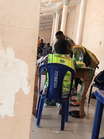The image depicts an indoor scene in Ajalli, Nigeria, characterized by light-colored walls showing extensive paint peeling on the left foreground. A person with dark braided hair is seated with their back to the viewer, wearing a lime green reflective safety vest bearing a logo and text. They occupy a blue plastic chair with "DD RENTALS" stenciled on the back, positioned at a rustic wooden table. Behind the seated individual, a male with short dark hair, wearing a grey and orange vest, leans over the table. To his right, another individual, partially visible, wears an orange reflective vest and green pants, also attending to the table. A plastic water bottle rests on the table surface. Black plastic bags are visible on the tiled floor beneath the table. A large reflective surface on the left wall captures a broader view of the environment, showing multiple structural pillars with grey capitals, a ceiling equipped with several white ceiling fans, and additional individuals standing and seated in the background, suggesting a waiting area or public gathering.