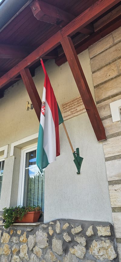 The image shows a Hungarian flag mounted on the side of a building. The building appears to be made of light-colored stucco and has a stone foundation. There is a window to the left of the flag, and a planter box with flowers sits on the windowsill. A plaque is mounted on the wall to the right of the flag, containing Hungarian text. The flag is attached to a wooden pole, which is held by a green metal bracket. The building has a wooden overhang with red beams. The scene is outdoors, with what appears to be trees reflected in the window. The text on the plaque is a dedication to the building's construction, indicating it was built between 2004 and 2005. The location is identified as Cserszegtomaj, Hungary.