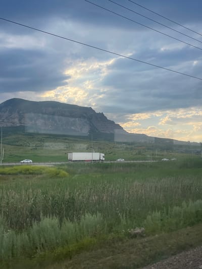 A wide shot captures a mountain range under a cloudy sky with sunbeams breaking through, located in or near Mineralnye Vody, Russia. The foreground is dominated by a field of green grass and vegetation. A highway runs horizontally across the middle of the frame, with several vehicles, including a white truck and two cars, traveling on it. A large, imposing mountain with rugged, rocky slopes rises in the background, its peak partially obscured by clouds. Overhead, power lines stretch across the sky. The lighting suggests it is either early morning or late afternoon, with soft sunlight illuminating parts of the clouds and the distant landscape.