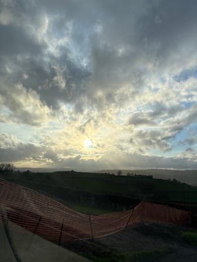The image depicts a landscape in Shrewsbury, United Kingdom under a sky filled with clouds and bright sunlight. An orange safety fence is visible on the right side, with a view of green fields and distant hills in the background. The sun is positioned behind the clouds, creating a diffused light effect.